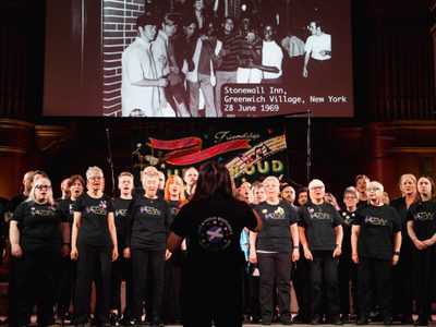 A conductor faces the Loud and Proud choir who are singing under a projection of a black and white picture outside the Stonewall Inn.