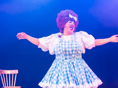 Johnny McKnight is dressed as a pantomime dame, with a red wig and blue and white gingham pinafore. He stands centre-frame and has his arms spread wide. The background is a deep sea blue.