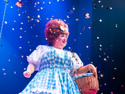 Johnny McKnight, a pantomime dame stands on stage holding a little wicker basket. Confetti falls from the sky.