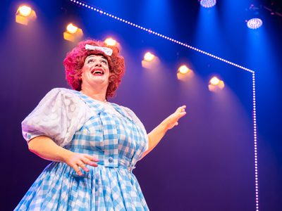 Johnny McKnight, a pantomime dame, stands on stage. He is looking to the lights with a awed and hopeful expression.