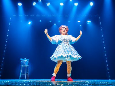 Johnny McKnight, a pantomime dame, is mid-dance. He wears red shoes and a blue and white gingham pinafore. The lights are on him and a small wooden chair sits at the back of the stage.