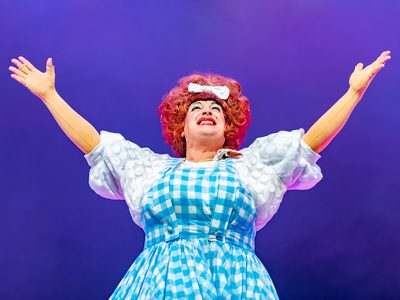 Johnny McKnight, a pantomime dame, stands on stage with a jubilant smile and arms raised to the sky. It is a triumphant pose. The background is purple.
