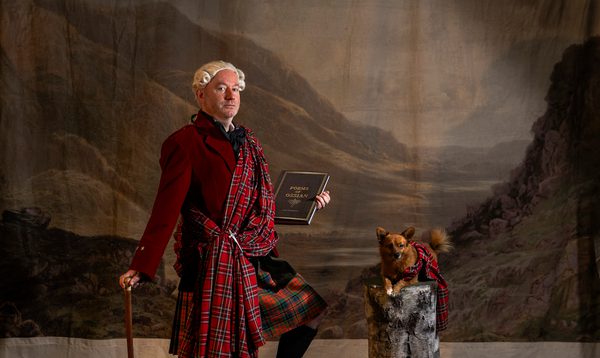actor Martin O'Connor stands in front of a Scottish landscape, wearing a great kilt and holding the poems of Ossian, while a similarly outfitted small dog sits nearby.