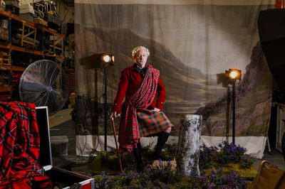 A man dressed in a kilt and Scottish garb stands in front of a painted backdrop and set of a Highland landscape. Behind him you can see a warehouse, fan and lights.