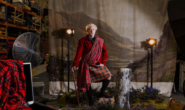 A man dressed in a kilt and Scottish garb stands in front of a painted backdrop and set of a Highland landscape. Behind him you can see a warehouse, fan and lights.