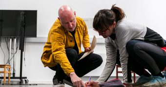 A person in a yellow jacket and another person in a grey top crouch on the floor over a large sheet of paper in a rehearsal space