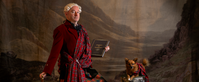 actor Martin O'Connor stands in front of a Scottish landscape, wearing a great kilt and holding the poems of Ossian, while a similarly outfitted small dog sits nearby.