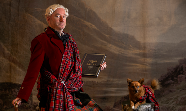 actor Martin O'Connor stands in front of a Scottish landscape, wearing a great kilt and holding the poems of Ossian, while a similarly outfitted small dog sits nearby.