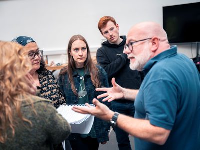 Director Dominic Hill talks to a small group of actors. They all watch him intently.