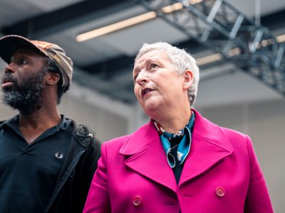 Blythe Duff and Jo Servi stand together. They are looking at something off-camera. Blythe wears a fuschia coat and Jo wears a black top and baseball cap.