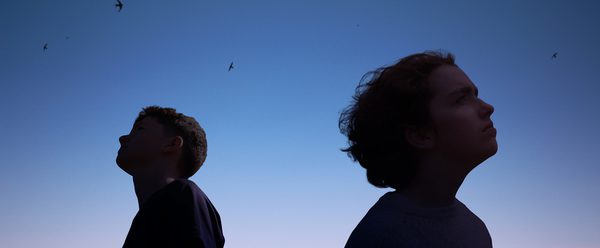 Two teenagers (a boy and a girl) stand almost back to back and they gaze at the dusk sky