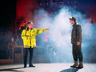 Actor Ewan Donald, wears a high-vis jacket. He points to fellow actor Lewis Fleming who is dressed as a soldier, the pair appear to be having a debate. There is white fog in the background.