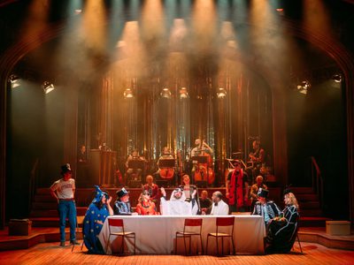 Several members of the ensemble sit at a long table - 'Last Supper' style. They are all dressed in Halloween costumes such as a ghost; wizard; and pumpkin.