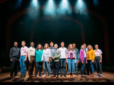 All fourteen members of the Small Acts of Love ensemble stand on stage they wear a variety of expressions from serious to sad to hopeful.