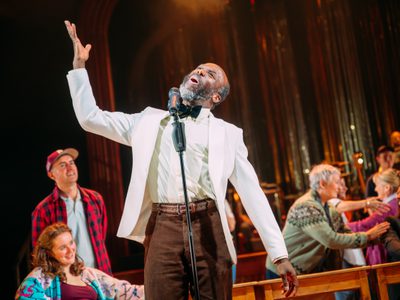 Jo Servi stands in the centre of the image, he wears a white suit jacket. He sings to a microphone and has his arm raised above his head - he is absorbed in his performance.