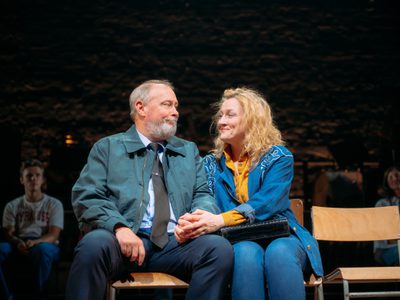 Barrie Hunter and Beth Marshall next to each other on wooden chairs. They look at each other with love and sadness and hold each others hand.