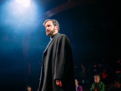 Actor Robbie Jack is dressed as a priest. He has a pensive expression. There is a bright soft blue light above him.