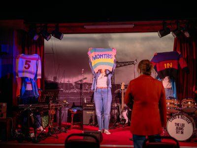 Three women stand on-stage. They hold up knitted jumpers with the words "5 Months In" sewn on them.