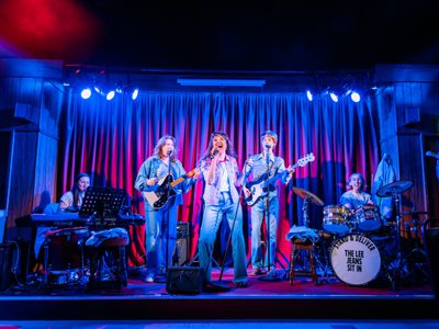 5 performers play as a band with a red curtained backdrop in 80s denim costume. The lighting is blue and white.