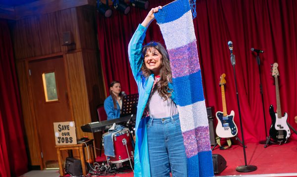 Chiara wears a cyan overcoat and jeans. She holds up a knitted scarf which is longer than her, she beams with determination.