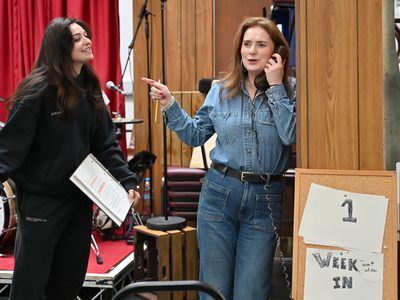 Jo Freer wears trendy denim and holds a telephone. She points to Chiara Sparkes who is trying to get her attention.