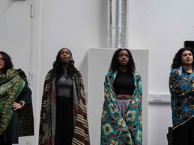 Hannah Lavery, Kimberley Mandindo, Chinyanta Kabaso and Jamie Marie Leary stand singing in a line. They're each wrapped in a coloured patterned cloth.