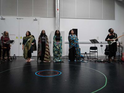 6 performers stand in a line. 1 looks down at a music stand, 4 are wrapped in colourful patterned fabric, 1 plays an electric guitar.
