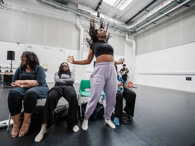 In a rehearsal Hannah Lavery, Kimberley Mandindo and Jamie Marie Leary sit in a line looking melancholy. Chinyanta Kabaso stands between them her head thrown back, arms thrown out.