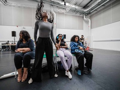 In a rehearsal Hannah Lavery, Chinyanta Kabaso and Jamie Marie Leary sit in a line looking melancholy. Kimberley Mandindo stands between them her head thrown back.