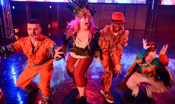 Four performers in vibrant, theatrical costumes crouch and reach toward the camera on a brightly lit stage, with colourful lighting, a reflective floor, and screens in the background.