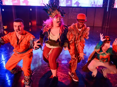 Four performers in vibrant, theatrical costumes crouch and reach toward the camera on a brightly lit stage, with colourful lighting, a reflective floor, and screens in the background.
