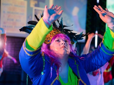 A young person stands in a bright light wearing a bright costume and feathered headdress. Their hands are raised.