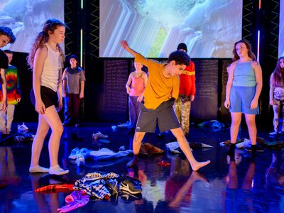 A group of young people stand forlornly on stage surrounded by cast off costumes. 1 young person dances in the middle.