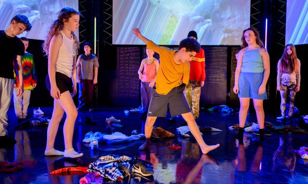 A group of young people stand forlornly on stage surrounded by cast off costumes. 1 young person dances in the middle.