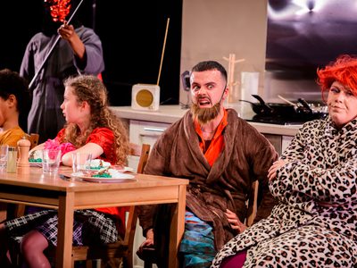 A group of young people sit roung a kitchen table. 1 has a disapproving look wears a leopard print dressing gown and a red wig. Another has an angry expression and wears a brown dressing gown and fake beard.