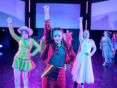 3 young people in cosplay costumes dance with one fist in the air. The lighting is pink.
