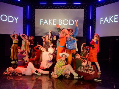 A group of young people in cosplay costumes create a theatrical tableaux with the words FAKE BODY projected on screens behind them. The lighting is warm.
