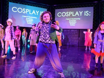 A group of young people stand on stage in Cosplay costumes. The lighting is blue and pink.