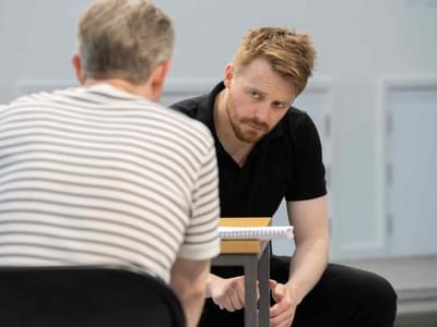 Martin and Jack sitting at a table in the rehearsal room. Martin has his back to the camera and Jack is watching him warily.