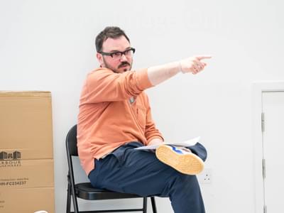 The director sits on a chair in the rehearsal room. He is pointing past the camera.