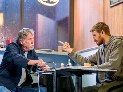 In a cafe set, Jack Lowden points firmly at Sean Gilder who sits across from him looking curious.