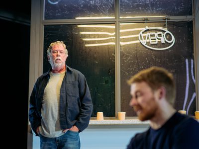 Sean Gilder stands looking seriously down at Jack Lowden in front of a window sill with used coffee cups on it. Strip lighting from above reflects in the window.