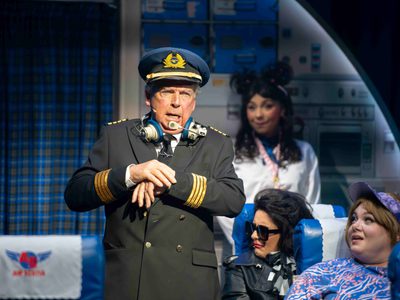 An airline captain stands in a plane aisle wearing his uniform. Passengers watch him.
