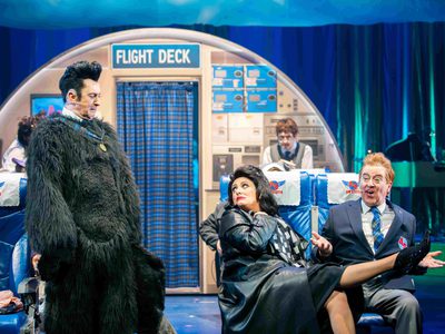 A man and woman sit together on a plane. The woman has her leg on the man's lap. A man in a hairy suit watches them in disgust.