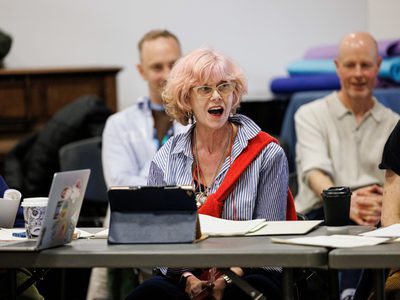 An actress with pink hair sits at a table her mouth is open in surprise two men sit behind her giggling.