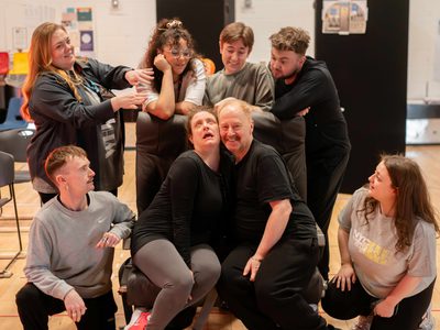 Two acotrs sit together they seem very keen on each other. More actors surround them.