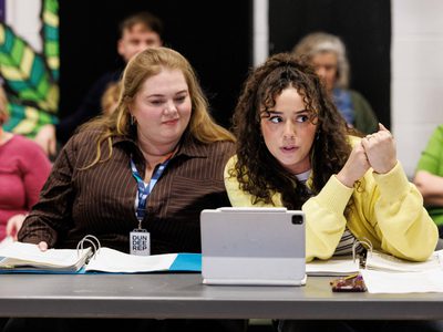 Two actresses sit at a table together reading a script,
