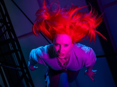 Vee Smith balances at the top of a pole, her hands are to her side. The lighting makes her hair look neon red and her body purple.
