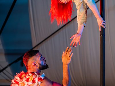 Vee Smith hangs down a pole, she reaches out to Sadiq Ali, who is crouching at the bottom of it. He also reaches for Vee. Sadiq has feathers on his back.