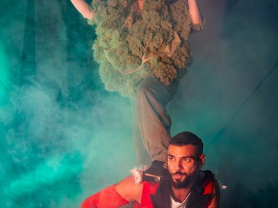 Sadiq Ali lies on the floor looking fed-up, he is wearing a red waistcoat. Vee stands on him, she is wearing a hood that looks like moss. She stares intently at something off-camera.